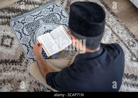 Muslimischer Mann liest den Koran, während er auf einer Gebetsmatte sitzt Stockfoto