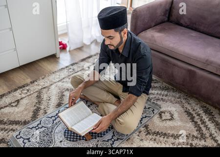 Muslimischer Mann liest den Koran, während er auf einer Gebetsmatte sitzt Stockfoto
