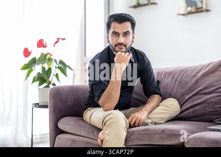 Bärtiger Mann, der auf einem Sofa im Wohnzimmer seines Hauses sitzt und in die Kamera blickt Stockfoto