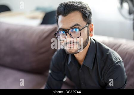 Bärtiger Mann, der auf einem Sofa im Wohnzimmer seines Hauses sitzt und in die Kamera blickt Stockfoto