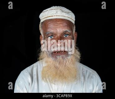 Mirsharai, Bangladesch – 20. September 2020: Blick auf einen älteren Mann mit markanten blauen Augen, einem verwitterten Gesicht und einem langen, hellen Bart, mit einer weißen Mütze, vor einem schwarzen Hintergrund. Stockfoto