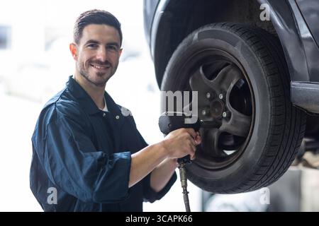 Männlicher Mechaniker, der neben dem angehobenen Autorad steht, entfernt die Radmuttern mit einem Druckluftschlüssel in der Garage Stockfoto