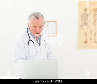 Männlicher Arzt mit weißem Mantel mit Laptop im Büro mit Stethoskop, Zertifikat, Skelettdiagramm Stockfoto