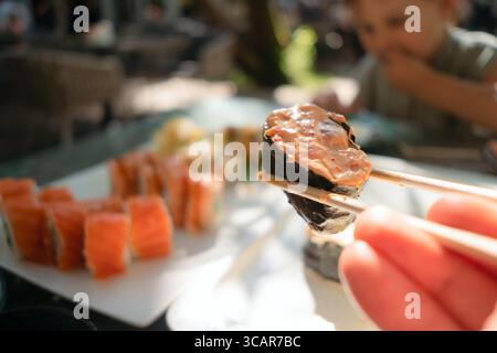 Nahaufnahme von frischem Lachs-Sushi mit Essstäbchen, authentisch japanischem Restaurant mit einem Kind, das im Hintergrund eine Mahlzeit genießt. Stockfoto