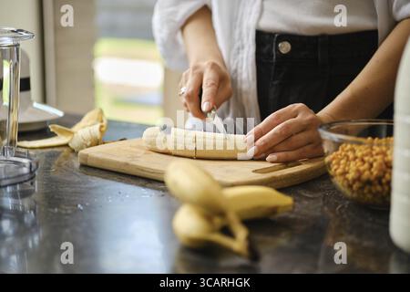 In einer warmen Küche schneidet eine Person reife Bananen auf einem hölzernen Schneidebrett. Frische Sanddornbeeren und andere Zutaten sind sichtbar und zeigen t Stockfoto