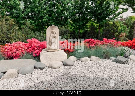 Ruhiger japanischer Garten mit Steinstatue, lebhaften Azaleen und friedlicher Felsenlandschaft im Frühling. Stockfoto