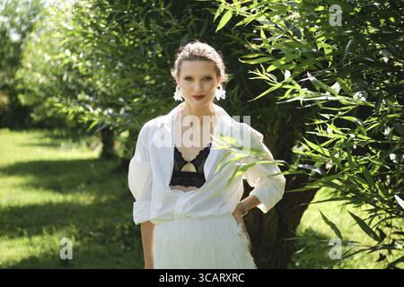 Ein Model posiert selbstbewusst in einem üppigen Garten und trägt ein stilvolles Outfit mit schwarzem Bralette und leuchtendem weißem Rock. Sonnenlicht filtert durch GREE Stockfoto