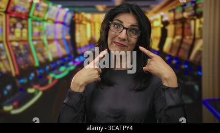 hispanische Frau mittleren Alters zeigt Finger auf Wangen neben leuchtenden Spielautomaten-Reihen im Casino; Freude. Stockfoto