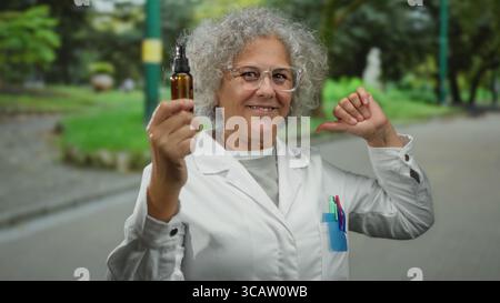 Ältere Frau mit lockigen Haaren in einem Labormantel, die stolz eine kleine Flasche auf einer Stadtstraße hält, umgeben von Grün und urbanen Details, die sich selbst zeigen Stockfoto