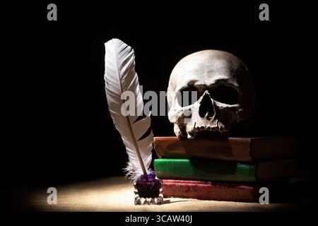 Drama oder Theater- und Literaturkonzept. Altes Tintenständer mit Feder in der Nähe des Schädels auf Büchern vor schwarzem Hintergrund. Dramatisches Licht. Stockfoto