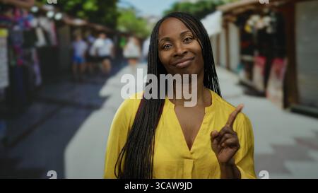 Schwarze Frau mit gelbem Hemd mit lockigen Haaren steht draußen in einer lebhaften Marktlage. Stockfoto