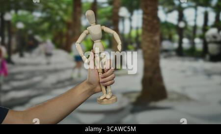 Hand hält eine Holzpuppe auf einer Stadtstraße mit verschwommenen Figuren im Hintergrund, die den Alltag in einem städtischen Gebiet darstellen. Stockfoto