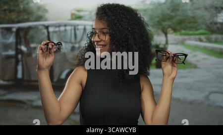 Junge Frau mit lockigem Haar im Freien, die zwei Brillen in jeder Hand hält und neugierig und besinnlich in einer grünen Parklandschaft aussieht Stockfoto