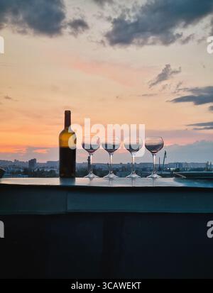 Flasche Wein und vier Gläser auf dem Dach bei Sonnenuntergang, Dämmerungshimmel und urbane Landschaft im Hintergrund. Stockfoto