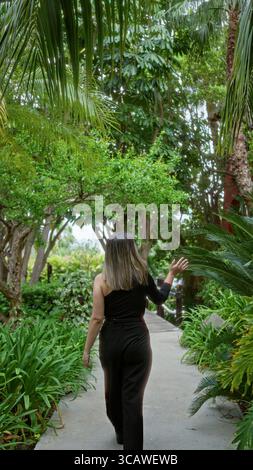 Frau schlendert auf einem üppigen tropischen Hotelweg, umgeben von lebhaftem Grün in einer ruhigen Resortlage unter einem Baldachin aus Palmblättern, die eine tr schaffen Stockfoto