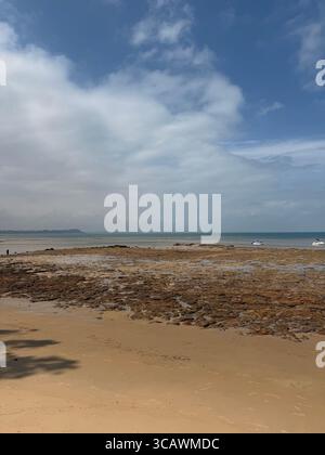 Sandstrand mit felsiger Küste unter teilweise bewölktem Himmel, Ebbe und weit entfernten Booten am Horizont Stockfoto