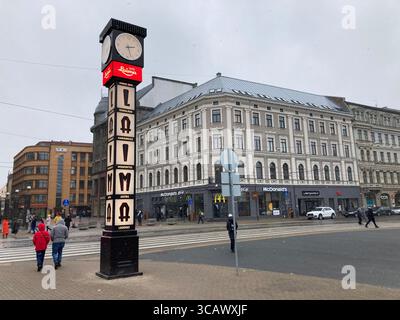 Die ikonische Laima-Uhr in Riga, eingerahmt von Schnee, mit ihrem historischen Charme und ihrem melodiösen Gesicht, das sanft vor einer Wintersilhouette leuchtet. Stockfoto