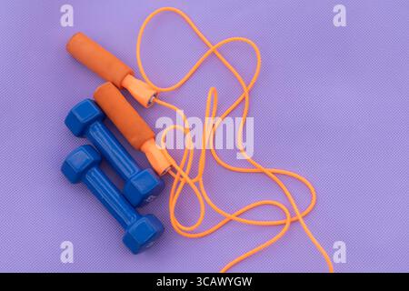 Ein Paar blaue Kurzhanteln und ein orangenes Sprungseil werden auf einer violetten Oberfläche platziert. Stockfoto