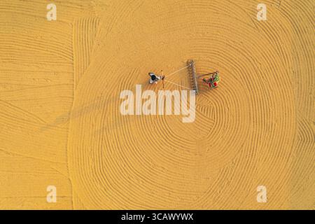 Bogura, Bangladesch - 24. Dezember 2018: Aus der Luft sehen zwei Arbeiter, winzig auf der riesigen ockerfarbenen Leinwand, die den Boden in konzentrischen Kreisen sorgfältig bearbeiten, ihre Schatten erstrecken sich lang und schlank. Stockfoto