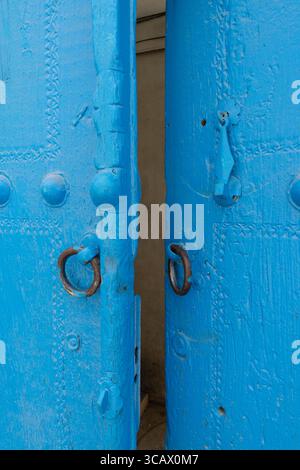 Eine Nahaufnahme von zwei leuchtend blauen, verwitterten Holztüren, die leicht offen stehen und mit einem detaillierten Metallklopfer und Ringgriffen verziert sind. Stockfoto