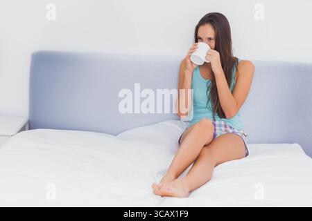 Frau mit türkisfarbenem Oberteil schlürft aus einem weißen Becher, während sie auf dem Bett im Schlafzimmer sitzt, Kopierraum Stockfoto