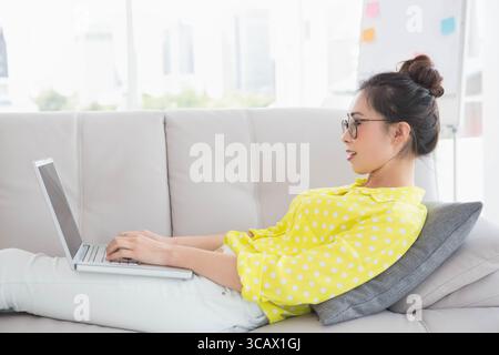 Chinesische Frau, die sich auf dem grauen Sofa liegend und auf dem Laptop neben dem Flipchart im Wohnzimmer, Kopierraum geschrieben hat Stockfoto