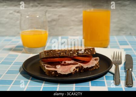 Vollkornsandwich mit Schinken und Tomatenscheiben, serviert auf einem schwarzen Teller mit Orangensaft, bereit für ein nahrhaftes Frühstück Stockfoto
