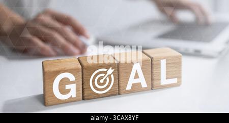 Ein Geschäftsmann arbeitet fleißig an seinem Laptop, mit Holzblöcken neben ihm, die „ZIEL“ schreiben. Der Buchstabe „O“ wird kreativ durch ein Zielsymbol, s, ersetzt Stockfoto