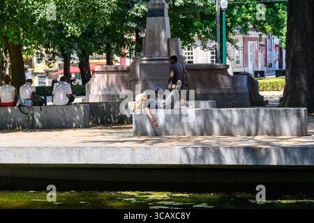 23/07/2025 Porto, Portugal. Eine junge Frau, die sich an einem Brunnen im Jardim da Cordoaria mit Buch und Musik entspannt. Mit Jeans, T-Shirt und Baseballmütze. Foto: © Simon Grosset Stockfoto
