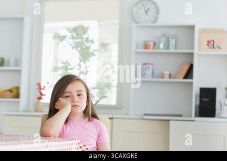 Kind Mädchen sitzt am karierten Tisch in der Küche unter der Wanduhr, blickt nach draußen, Kopierraum Stockfoto