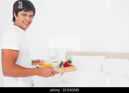 Mann, der neben dem Bett stand, hielt ein Holztablett mit Saft, Müsli und Rose im Schlafzimmer, Kopierraum Stockfoto