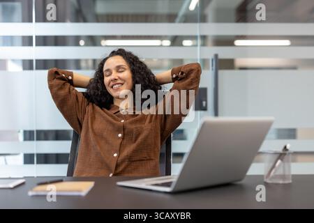 Büroangestellte lehnte sich entspannt und lächelnd auf ihrem Arbeitsplatz in einem Stuhl zurück. Erfasst einen Moment der Zufriedenheit und Leichtigkeit am Arbeitsplatz und veranschaulicht die Vereinbarkeit von Beruf und Privatleben und die positive Einstellung. Stockfoto