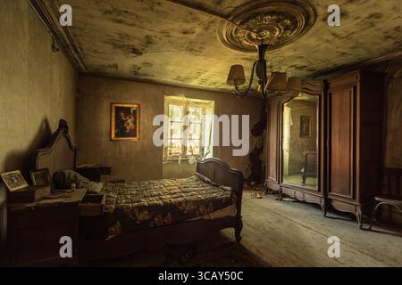 Verlassenes Vintage-Schlafzimmer mit antiken Möbeln, abblätternden Wänden und natürlichem Licht durch kaputte Fenster in einem vergessenen alten Haus Stockfoto