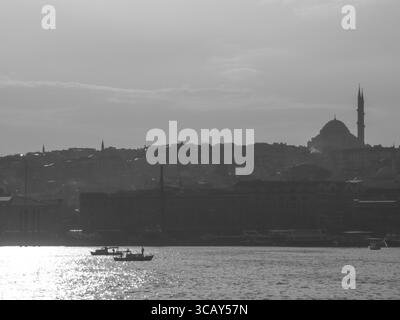 Schwarz-weiße Silhouette der Skyline von Istanbul mit Moschee und Booten auf glitzerndem Wasser bei Sonnenuntergang Stockfoto