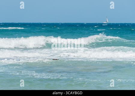 Malerischer Blick auf die Wellen, die unter einem klaren blauen Himmel an die Küste stürzen, mit einem Segelboot am Horizont. Eine ruhige Meereslandschaft Stockfoto