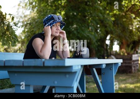 Eine schöne Frau, die draußen an einem Picknicktisch sitzt, einen sonnigen Tag genießt und einen Eimerhut trägt. Frau mittleren Alters, die im Schatten eines Baumes ruht Stockfoto