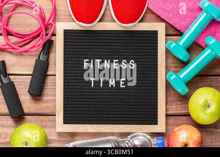 Fitnesszeit und Sportkonzept. Rote Turnschuhe, Äpfel, Sprungseil, Kurzhanteln, rosafarbenes Handtuch und schwarzes Schriftfeld mit den Worten Fitness Time auf hölzernem Hintergrund Stockfoto