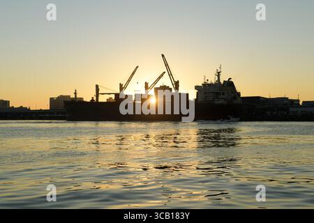 Schöne Silhouette von Kränen, die Containerfrachtschiff bei Sonnenaufgang laden, Reflexion auf Wasser, Durban Hafen, KwaZulu-Natal, Südafrika, Transport Stockfoto