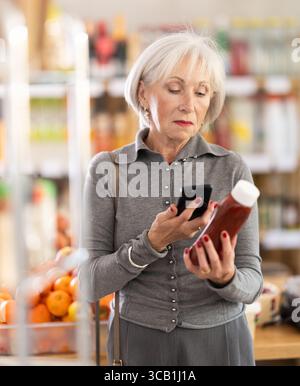 Ältere Käuferinnen scannen QR-Code auf Flaschenketchup-Etiketten in der Lebensmittelabteilung des Supermarktes Stockfoto