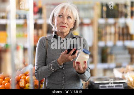Ältere Käuferinnen scannen QR-Code auf Flaschenmayonnaise-Etikett in der Lebensmittelabteilung des Supermarktes Stockfoto