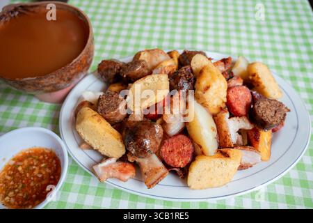 Nahaufnahme einer traditionellen kolumbianischen Picada mit Chorizo, Morcilla, Arepa, Schweinebauch und Kartoffeln auf einem Teller über einem grünen karierten Tisch Stockfoto