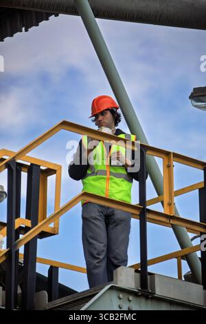 Arbeiter des Auftragnehmers kletterte mit Sicherheitsausrüstung und Schutzhelm mit dem Telefon und trug Arbeitskleidung auf eine industrielle Metallkonstruktion Stockfoto
