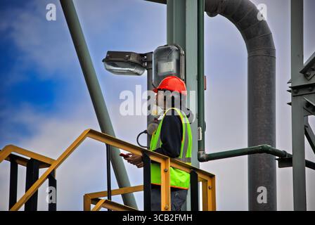 Arbeiter des Auftragnehmers kletterte mit Sicherheitsausrüstung und Schutzhelm mit dem Telefon und trug Arbeitskleidung auf eine industrielle Metallkonstruktion Stockfoto