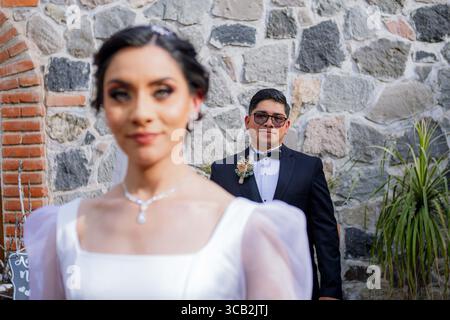 Vor einer rustikalen Steinmauer schaffen verschwommene Braut im Vordergrund und fokussierter Bräutigam im Hintergrund ein künstlerisches und elegantes Porträt. Stockfoto