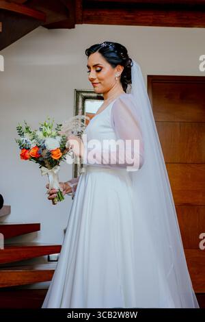 Ein Porträt einer lächelnden Braut drinnen, in ihrem weißen Kleid und mit einem wunderschönen Blumenstrauß. Das Bild ist intim und ruhig. Stockfoto