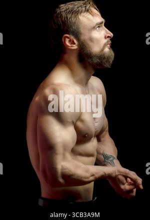 Ein muskulöser Mann mit Bart schaut hoch, sein Körper ist vor schwarzem Hintergrund beleuchtet. Stockfoto