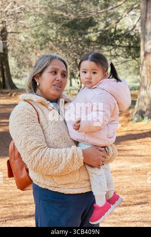 Mutter hält ihre Tochter in einem Herbstwald. Beide sind warm gekleidet; sie trägt eine pinkfarbene Jacke und Turnschuhe, er trägt eine beige Jacke. Ein warmes, vertrautes Bild Stockfoto