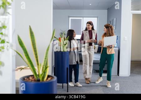 Verschiedene weibliche Kollegen, die an Pflanzgefäßen im Flur vorbeilaufen und Kaffeetassen mit Tablet und Laptop halten Stockfoto