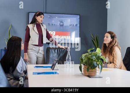 Verschiedene Kollegen verwenden ein Tablet an einem weißen Konferenztisch, während die Frau den Fortschritt auf dem Bildschirm vorstellt Stockfoto