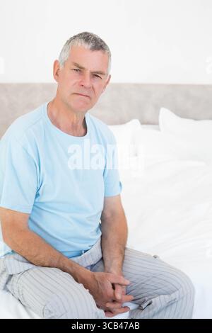 Älterer Mann, der im Schlafzimmer auf dem Bett sitzt, mit grauem Kopfteil, Hemd, gestreifte Hosen, die die Hände schließen Stockfoto